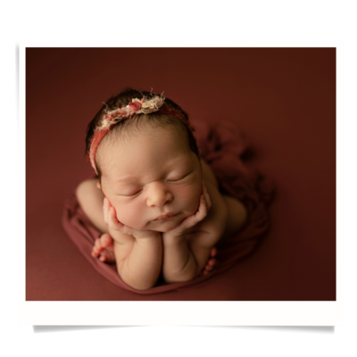 A baby in froggy pose on a mauve backdrop.