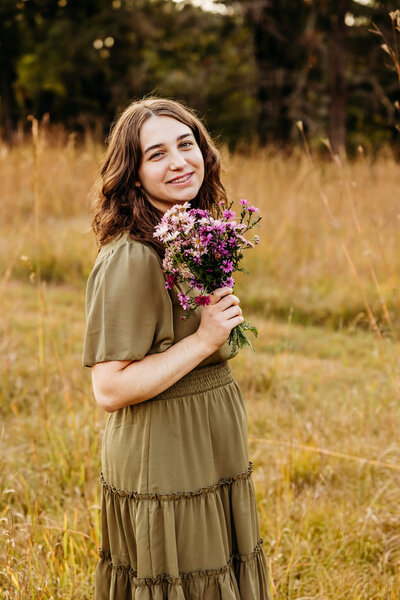 brown haired senior girl holding  a wildflower bouquet and smiling as she looks over  her shoulder in a field near Oshkosh, Wi