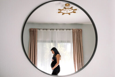 mirror reflection of pregnant woman  standing by window holding her baby bump