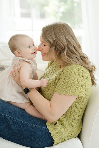 Mother holding her baby while laying on a white couch and leaning in to kiss her cheek while baby smiles