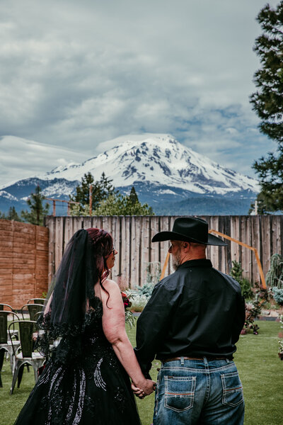 Bride in a black dress and groom in a cowboy hat hold hands in front of a wooden fence with a dramatic view of Mount Shasta in the background.