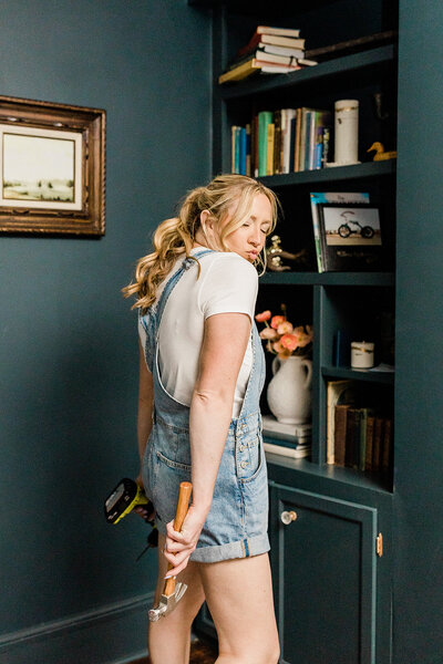A woman diyer carries a drill and a hammer making a funny face while wearing overalls in a room painted dark green.