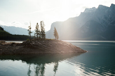 the sun rises over a calm Lake Minnewanka with mountains and trees reflected in the glacier-blue lake