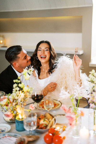 Bride sitting at table wearing fluffy jacket, groom sat next to her looking at here, table has food wine and florals design by Jessamine floral and events, New Jersey floral designer