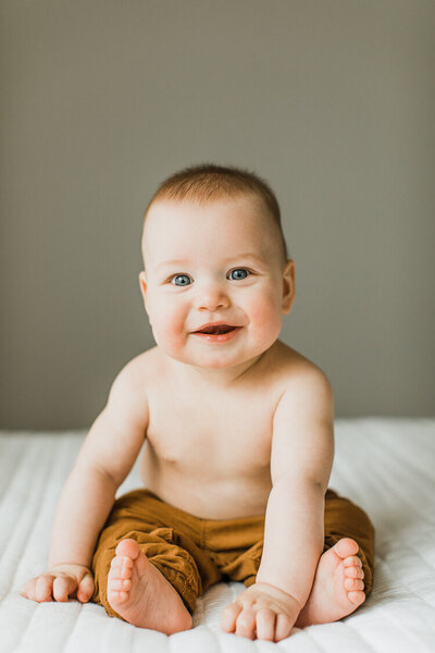 six month old baby boy sits on bed shirtless and smiles for photo