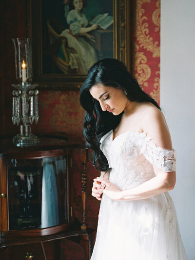 A bride with long black hair looks at her wedding ring in front of a window inside a historic mansion