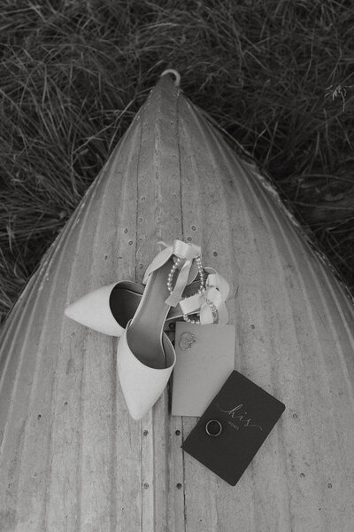 A black and white detail shot of shoes, vow books, and wedding rings all situated on a canoe.