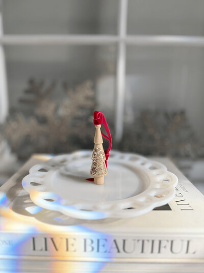 A small wooden holiday ornament with a red ribbon stands on a decorative white plate, which is placed on top of two stacked books titled “LIVE BEAUTIFUL” by Sweet Pea Ink Creative.