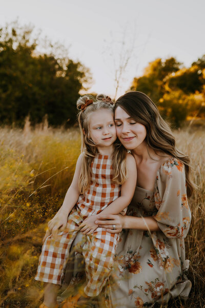 A mom and daughter in gingham plaid overalls sit together and cuddle in a tall, grassy field in fort worth, texas