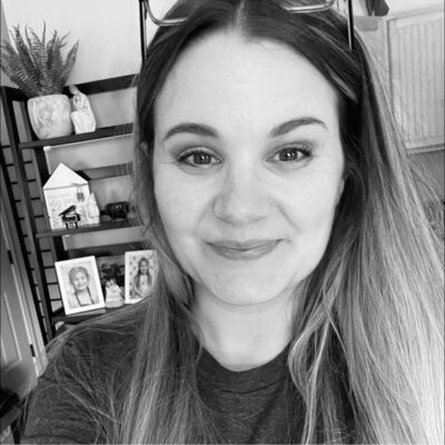 Black and white portrait of a smiling woman with long hair, wearing a dark top, in front of a bookshelf with home decor and framed photos.