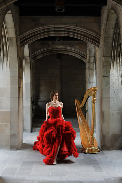 Light skinned woman with dark hair and long red ballgown is playing the harp. She is in an urban area, which is the Wrigley Building in Chicago