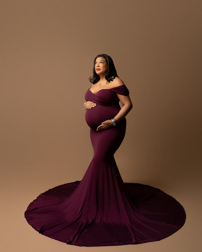 Studio maternity photo featuring deep plum fitted dress with off-the-shoulder neckline and dramatic silhouette