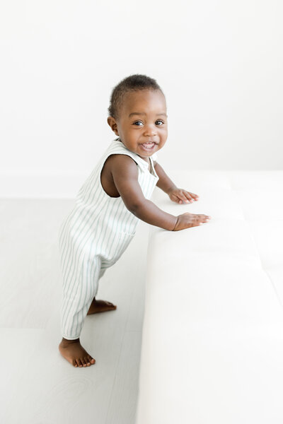 one year old baby boy smiles brightly as he stands wearing overalls during milestone portrait session