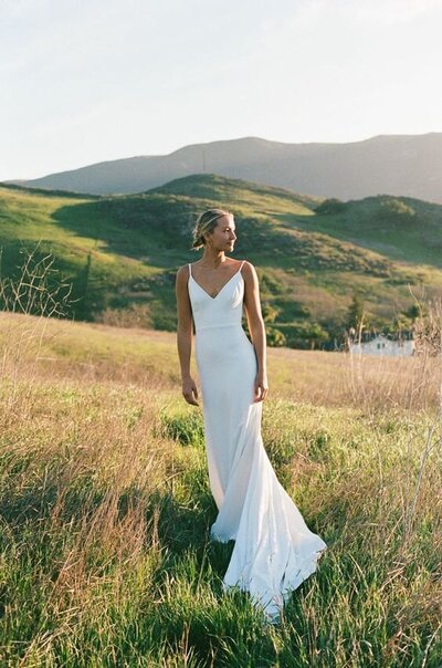 Bride standing in open field with hills behind her, wearing a simple white wedding dress