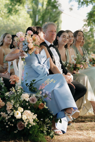 Photo of bride's parents and bridesmaids sitting in chairs during the ceremony and smiling