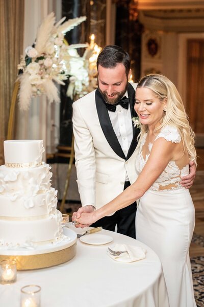 A bride and groom in elegant attire are smiling and cutting a three-tier wedding cake adorned with white decorative details. Candles and a floral arrangement are visible in the background, creating a festive atmosphere.