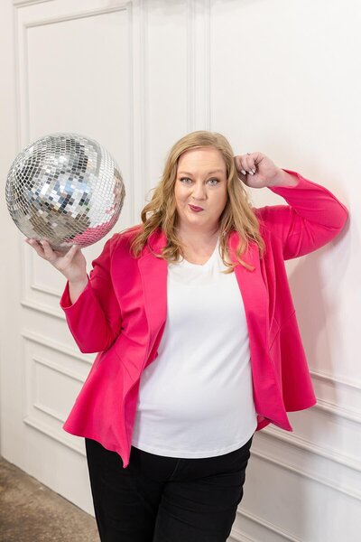 Confident woman in a hot pink blazer holding a disco ball during a personal branding photoshoot in a bright studio with white walls. Captured by a Seattle brand photographer, this playful and bold image reflects vibrant energy and creative expression for modern women entrepreneurs.