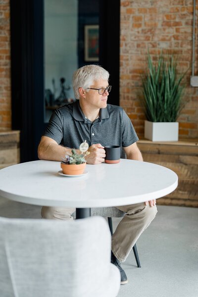 Columbus photographer captures brand photos of man sitting at a table while holding a mug and looking out the window in a studio space