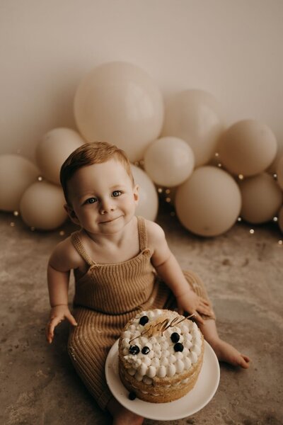 Adorable baby boy in brown romper with birthday cake and blueberries, seated in front of neutral balloon backdrop.