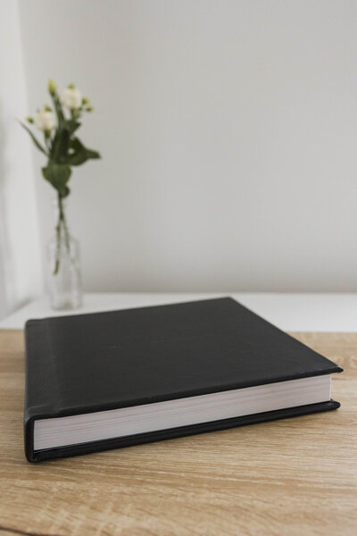 picture of a wedding hard coverd album with black leather cover on top of the table with a rose in the back.
