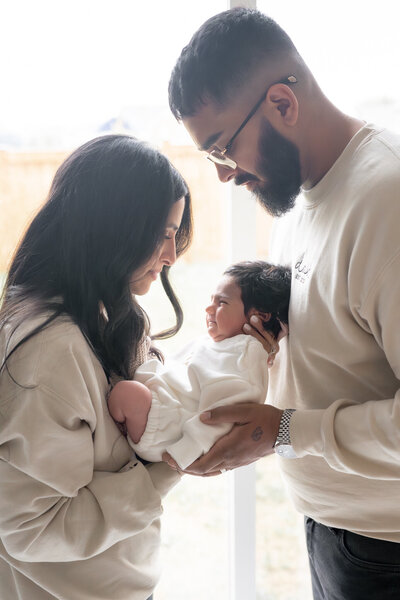 Lifestyle newborn photography session with parents holding baby in front of window with natural light— captured by Alliston and Barrie maternity, newborn, and family photographer.