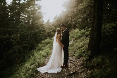 Bride and groom facing each and holding hands in a green forest as the sun sets.