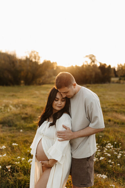 A pregnant mother wearing a white dress with a split in the front while her husband hugs her at sunset in a field of carrot weed in christchurch during their maternity photoshoot with lucy jane photography