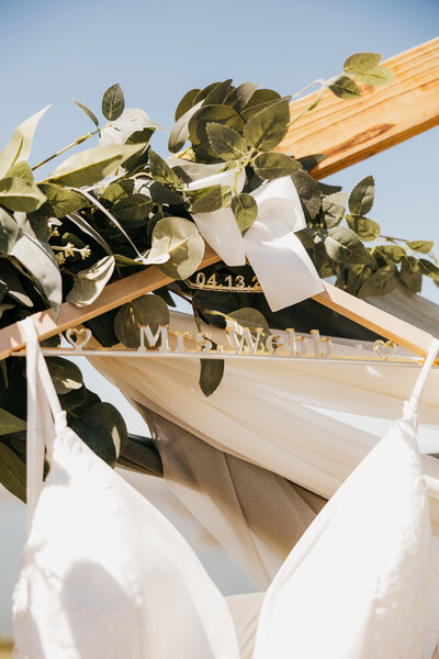 Details of the custom hanger holding the brides dress hanging on the arbor with greenery and light green draping