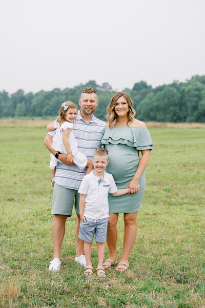 Family of four with pregnant mom smile at camera during summer photography session in Raleigh NC