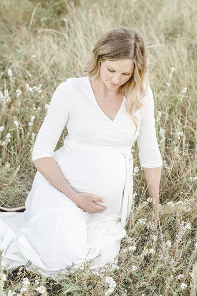 pregnant mother in white dress looking at belly while leaning on wall