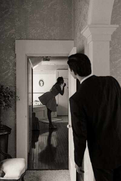 A bride puts on her lipstick in the mirror as groom watches through the doorframe.