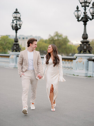 a man and woman in a white dress and tan suit walking towards the camera and looking at one another smiling and holding hands on the pont alexandre bridge in paris