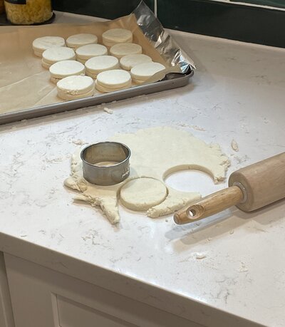 A hands-on baking scene showcasing fresh dough being rolled out on a floured white countertop. A metal cutter is used to shape perfectly round pastries, with a wooden rolling pin nearby. In the background, a metal tray holds neatly arranged dough circles, ready to be baked to perfection.