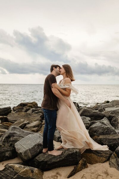 Beach portrait of a couple kissing on rocks, with ocean waves and cloudy sky in the background.