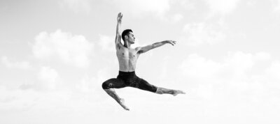 Black and white image of a shirtless male dancer performing an elegant mid-air leap against a cloudy sky, showcasing strength and grace.