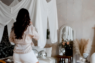 Emily Michele standing in a softly lit bedroom, adjusting her beige silk blouse — symbolizing mindful elegance, grounded confidence, and feminine embodiment.