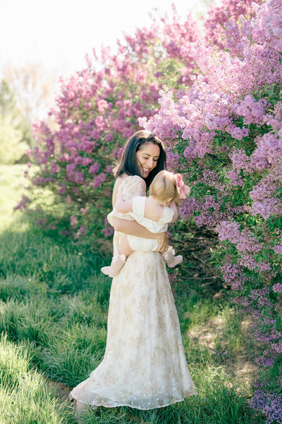 Colorado mom wearing a cream dress with her toddler daughter during a spring family photoshoot in front of purple lilac bushes in Erie, CO