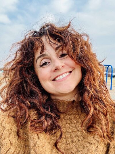  A woman with curly auburn hair smiles brightly while standing outdoors. She wears a cozy brown sweater, and the background features a playground with a cloudy sky.
