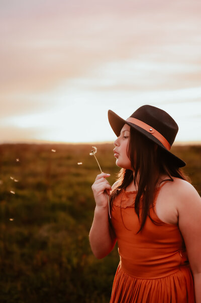 A woman wearing a brown floppy hat blowing a dandelion. 