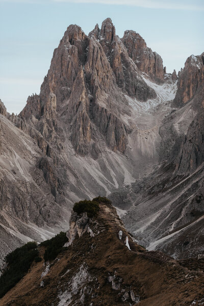 A groom helps his bride walk down a dirt hiking trail in the Dolomites. The bride wears a white dress with train and the mountain range rises behind them.