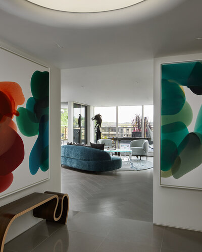 A foyer area with polished tile flooring, artistically curved bench, and colourful abstract wall art with an entryway to a living room with brown herringbone wood flooring, plush couches, and tall windows
