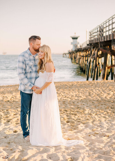 Husband kissing wifes forehead on the beach at maternity session Los Angeles by Ashley Nicole