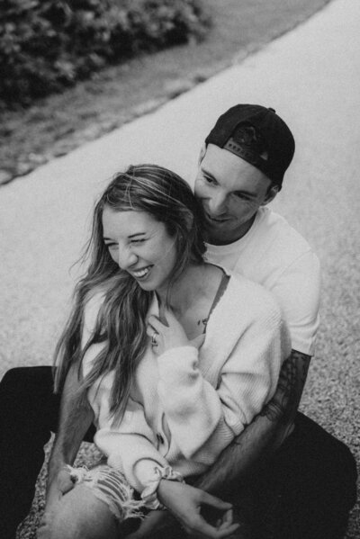 Dom sitting in her Jack's lap, her back against his chest, she is laughing and touching her hand to her chest as he whispers into her ear