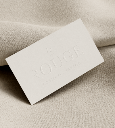 White business card with embossed text 'Le Rouge Cosmetic Tattoo' placed on top of neutral textile