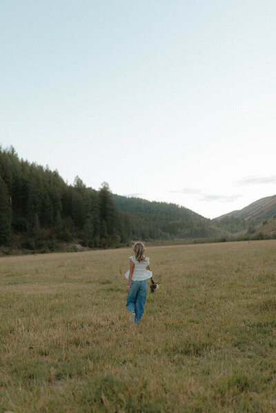 A high school graduate runs away from the camera towards trees and hills. She's holding a small bouquet of flowers in one hand.