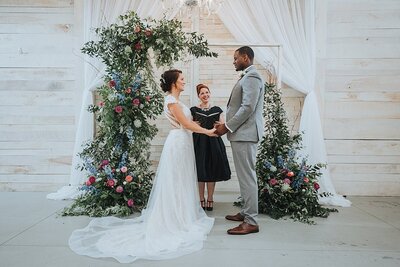 Mixed race bride and groom take their vows in front of a large lush two piece arbor full of greenery and pink and purple flowers inside a white wedding barn in Nashville