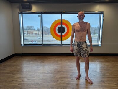 Before image of man posing shirtless in a gym with window and target background, used prior to professional dating photo session