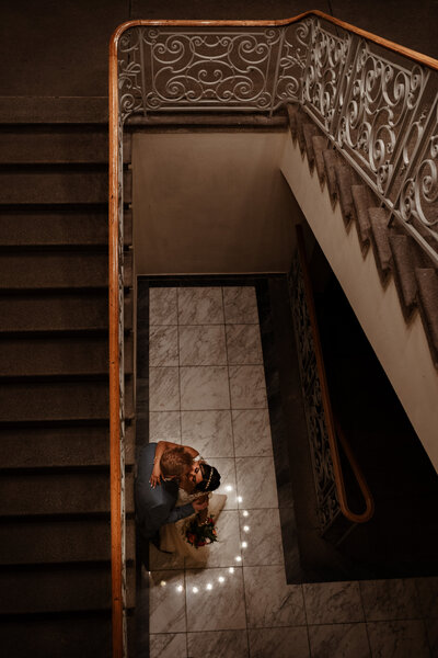 Brautpaar küsst sich am Fuße einer eleganten Treppe umrahmt von der Spiegelung des Kronleuchters im Boden.