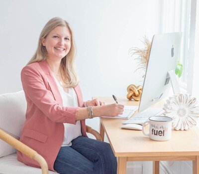 Maureen in pink blazer and jeans sitting at desk smiling at the camera