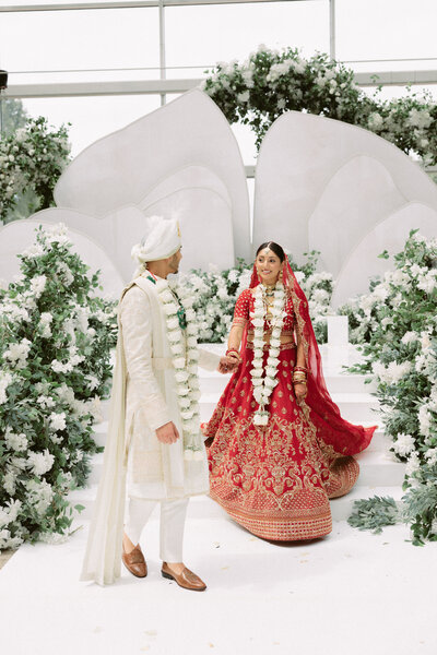 South Asian bride in her bright red lehenga walks with groom in his kurta to the aisle.